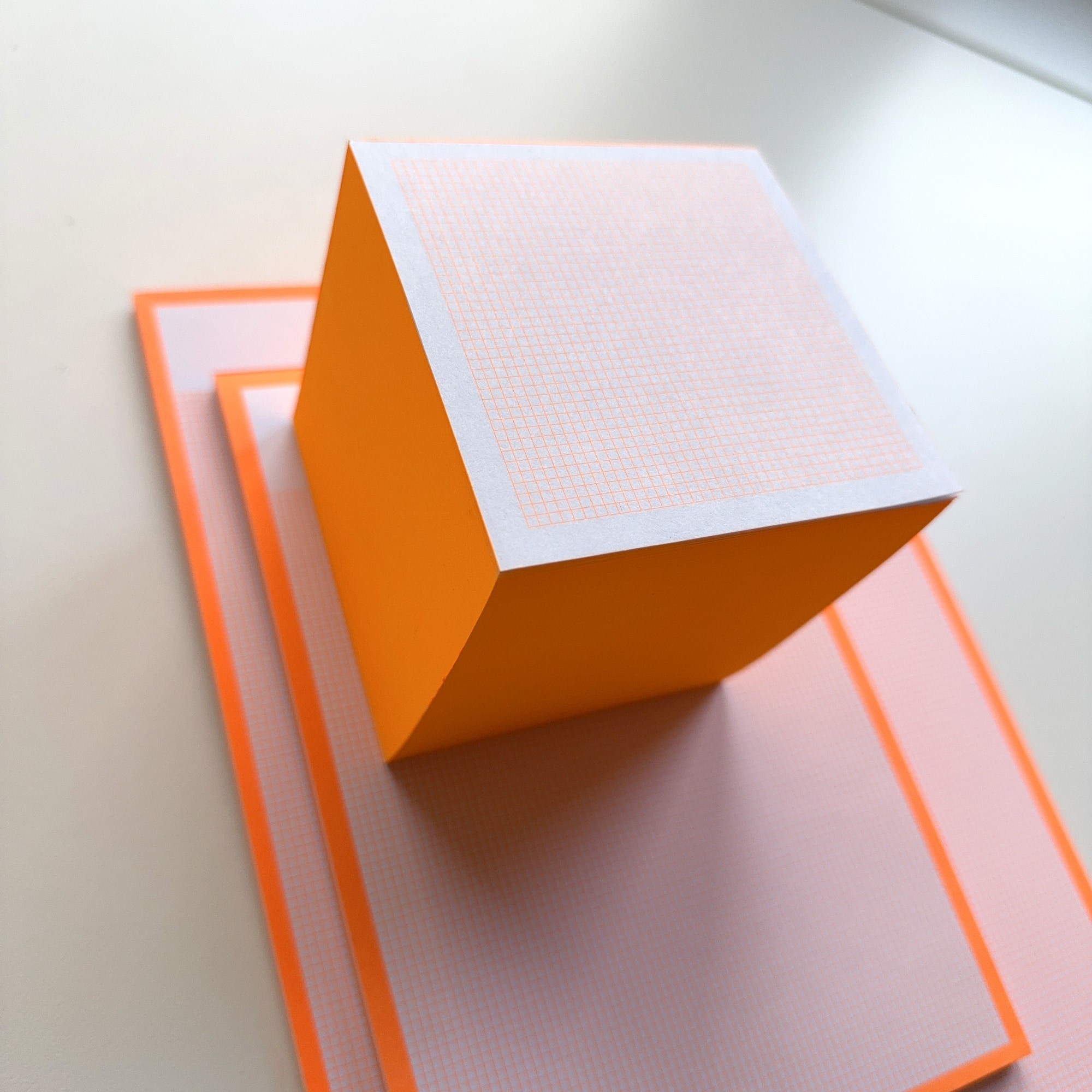 Notizklotz in Neonorange mit kariertem 2 × 2 mm Raster auf der Oberseite, 90 × 90 mm groß, leuchtend eingefärbte Seiten, aufgenommen vor weißem Hintergrund.